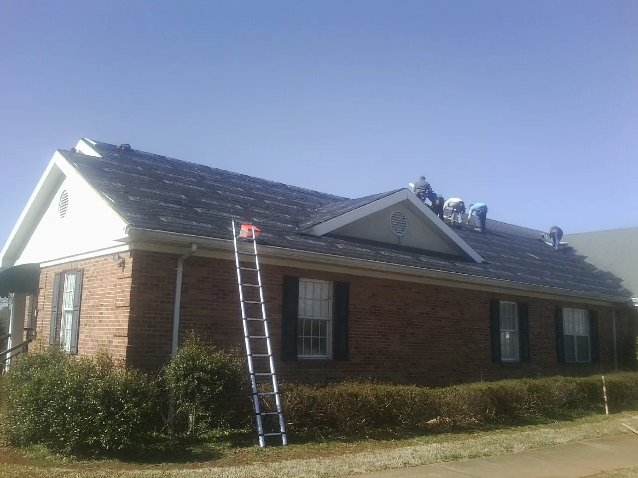 Crew tearing shingles off a brick ranch roof with a ladder set — Emergency Roof Repair work by our crew in Olar