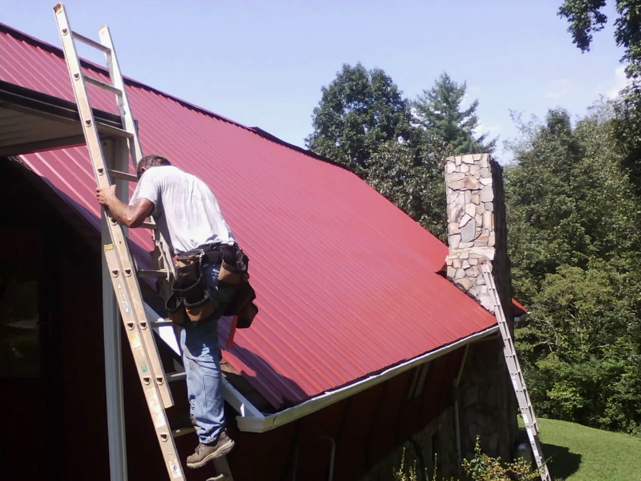 Roofer on a ladder beside a red metal roof and stone chimney — Soffit & Fascia Repair by Roofing Brothers in Olar